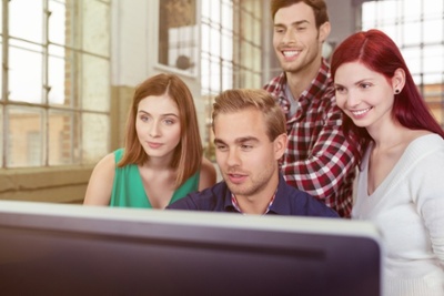 Informal young business team anxiously watching the success of their project as they stand grouped around a large screen desktop computer-1-1 Informal young business team anxiously watching the success of their project as they stand grouped around a large screen desktop computer-1-1